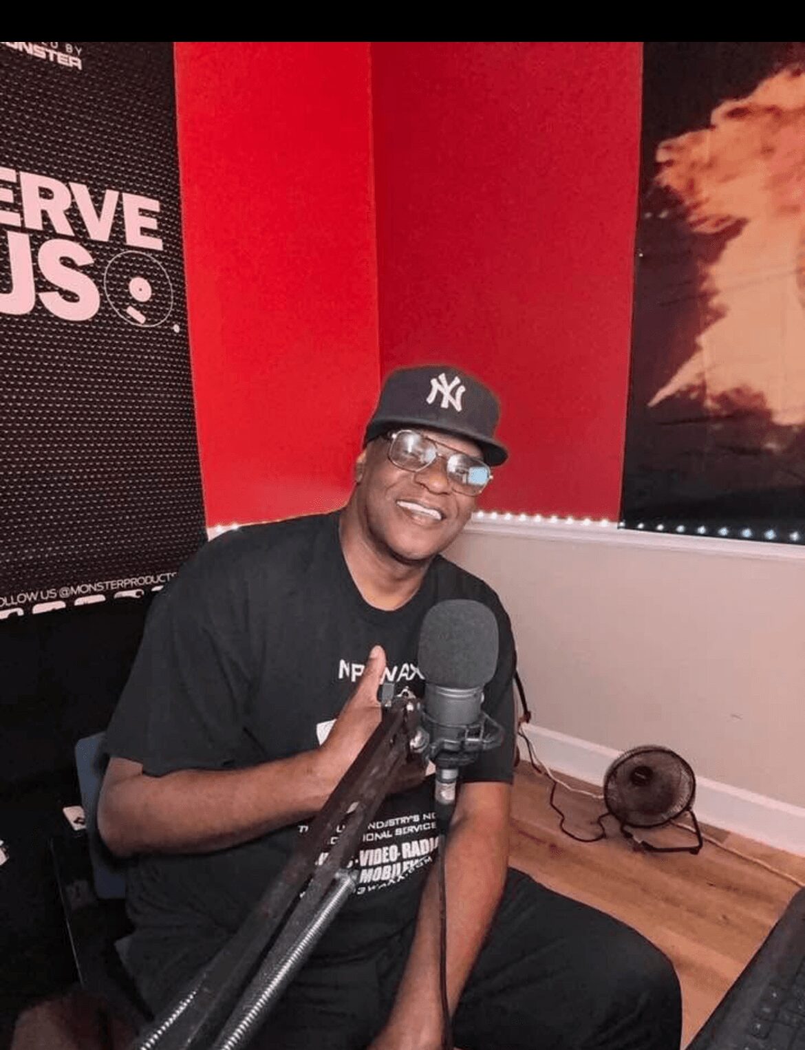 Person smiling, wearing glasses and a New York Yankees cap, sitting in a room with red and black walls, microphone in front.