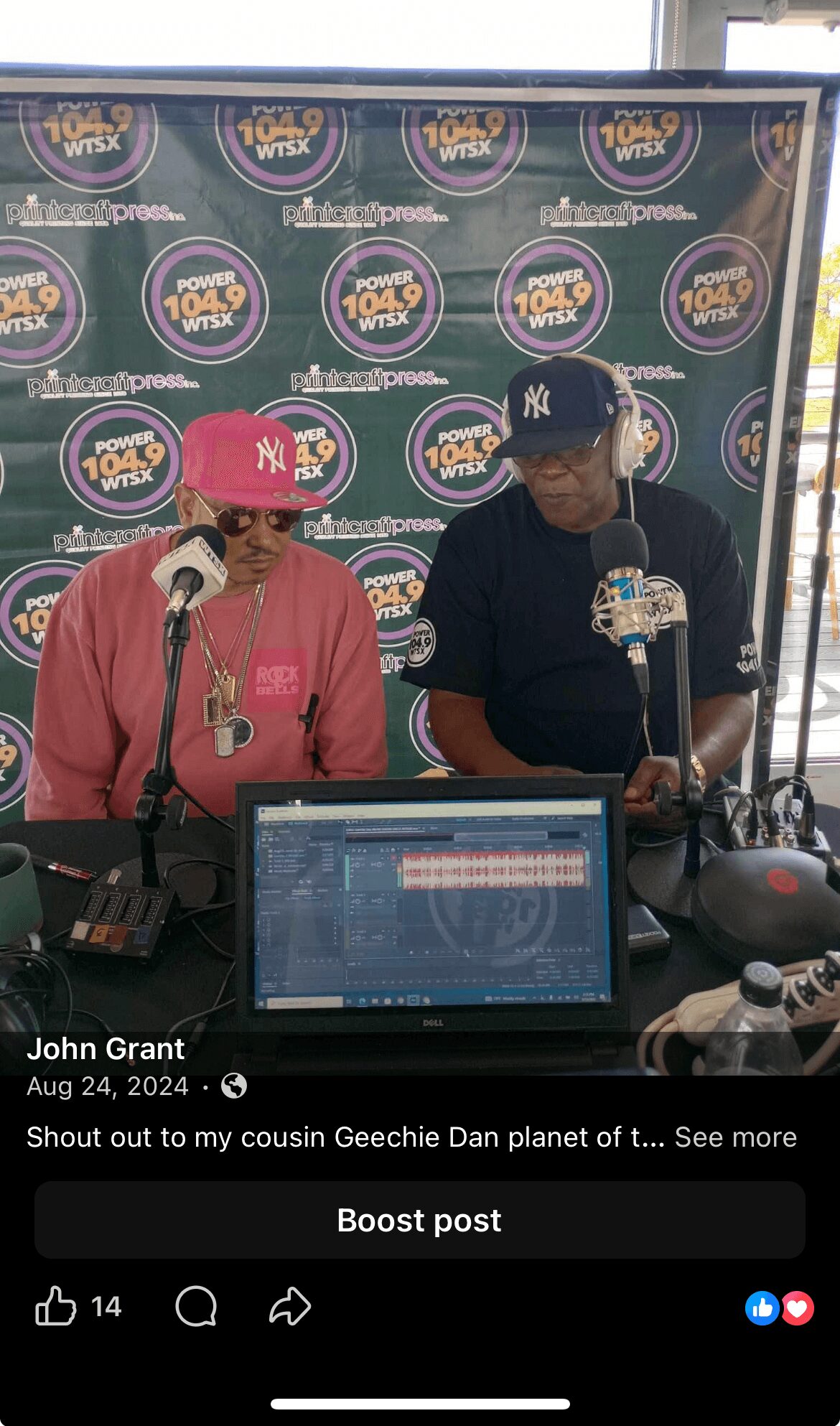 Two people sit at a table with microphones and a laptop, in front of a backdrop with logos, in a radio studio.
