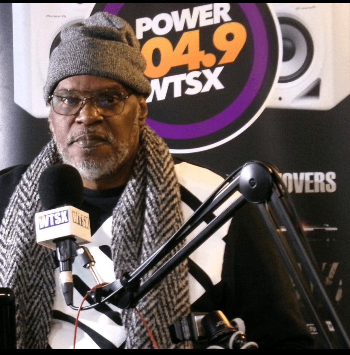 Man wearing glasses, a gray beanie, and a scarf speaking into a microphone at a radio station studio, with a Power 04.9 WTSX sign in the background.