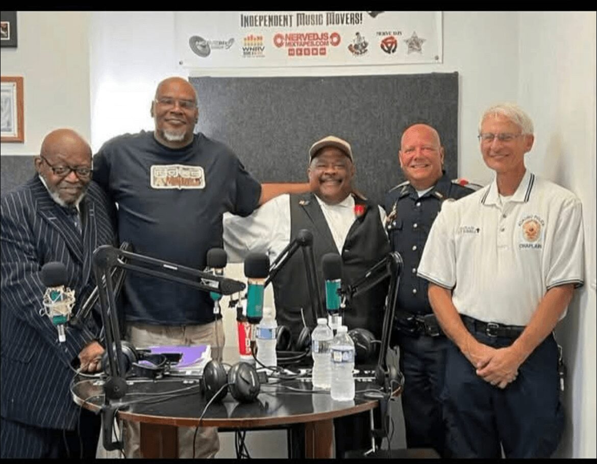 Five people standing in a room with microphones and recording equipment, smiling, with a banner in the background.