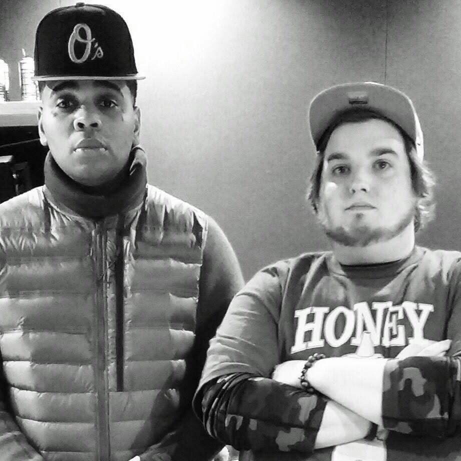 Two young men with caps, one wearing a puffer vest, standing indoors against a plain wall.