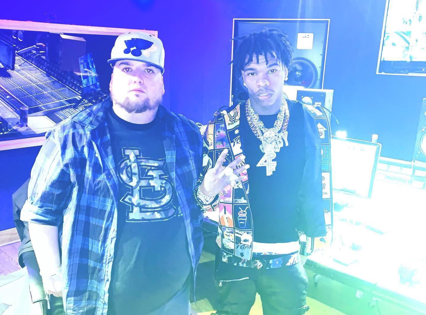 Two men standing in a room with electronic equipment, one wearing a plaid shirt and the other with jewelry and a patterned jacket.