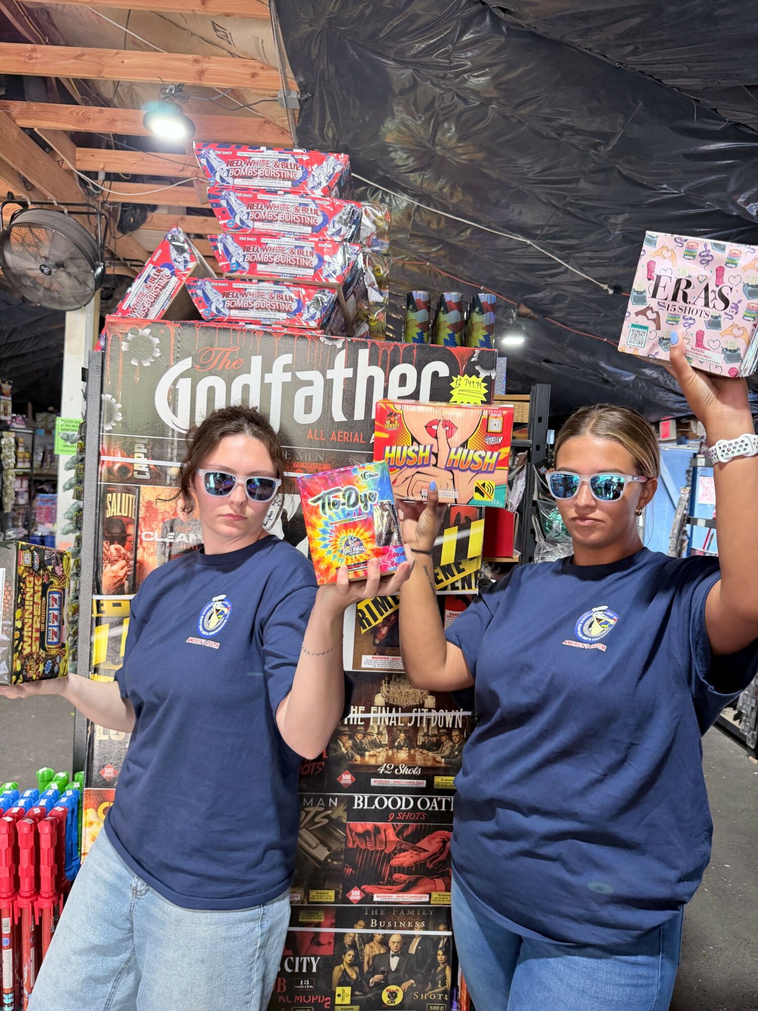 Two people wearing sunglasses and blue shirts stand in front of fireworks displays and colorful packaging at an outdoor event.