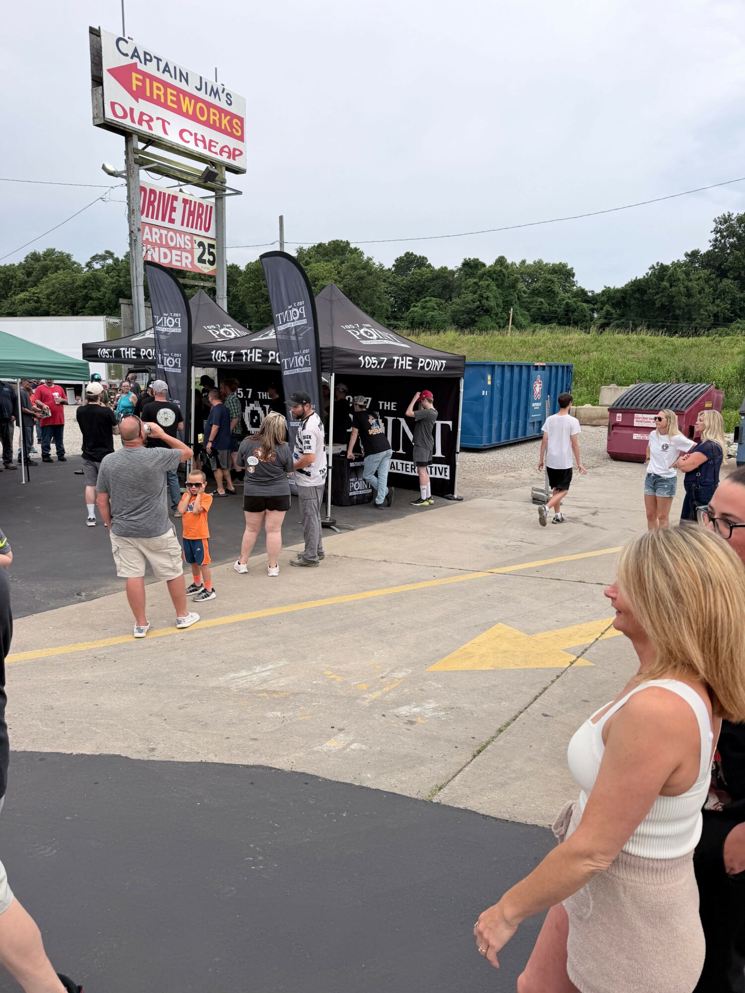People gather outside a store with a sign, some walking and others standing near a black tent, on a cloudy day.