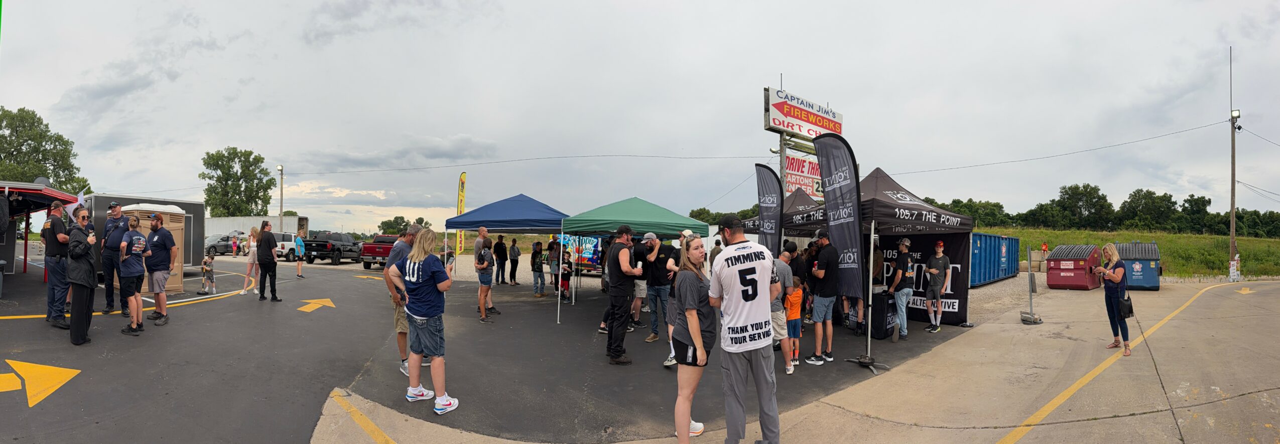 People gather outdoors near tents and a food truck, with cars parked in the background, under a cloudy sky.