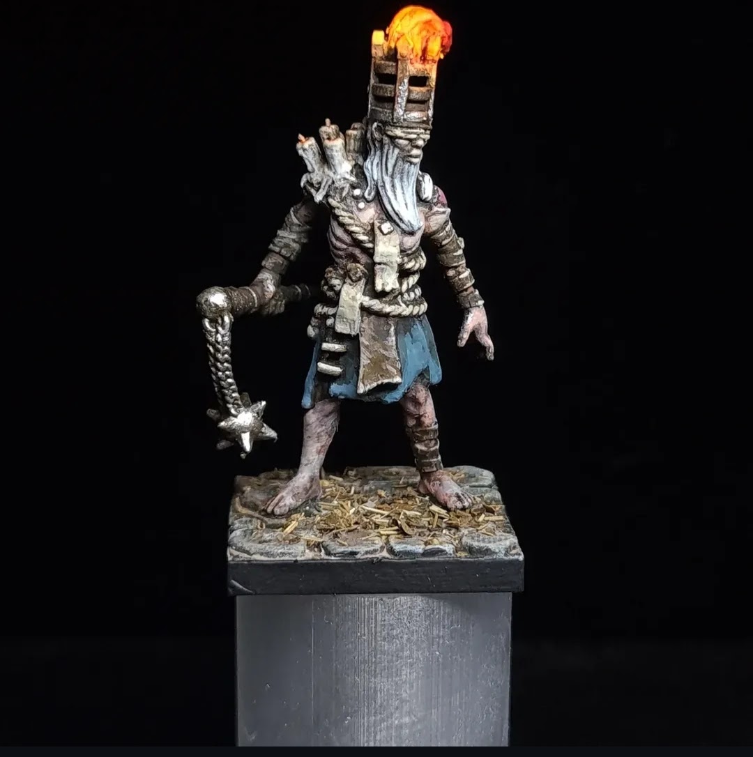 A detailed sculpture of a figure with a tall, flaming head, holding a chain and an object, standing on a textured base.