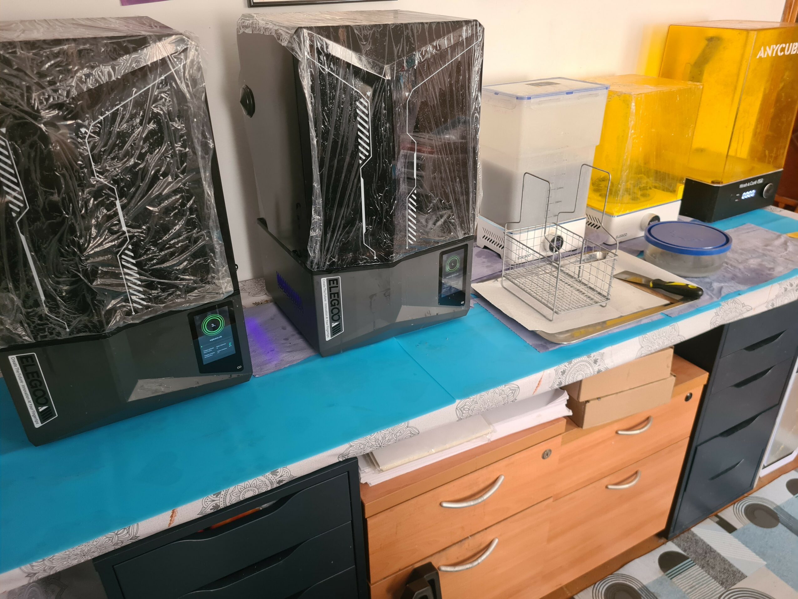 Three black electronic devices with plastic wrapping on a table, with yellow and clear containers and a notepad nearby.
