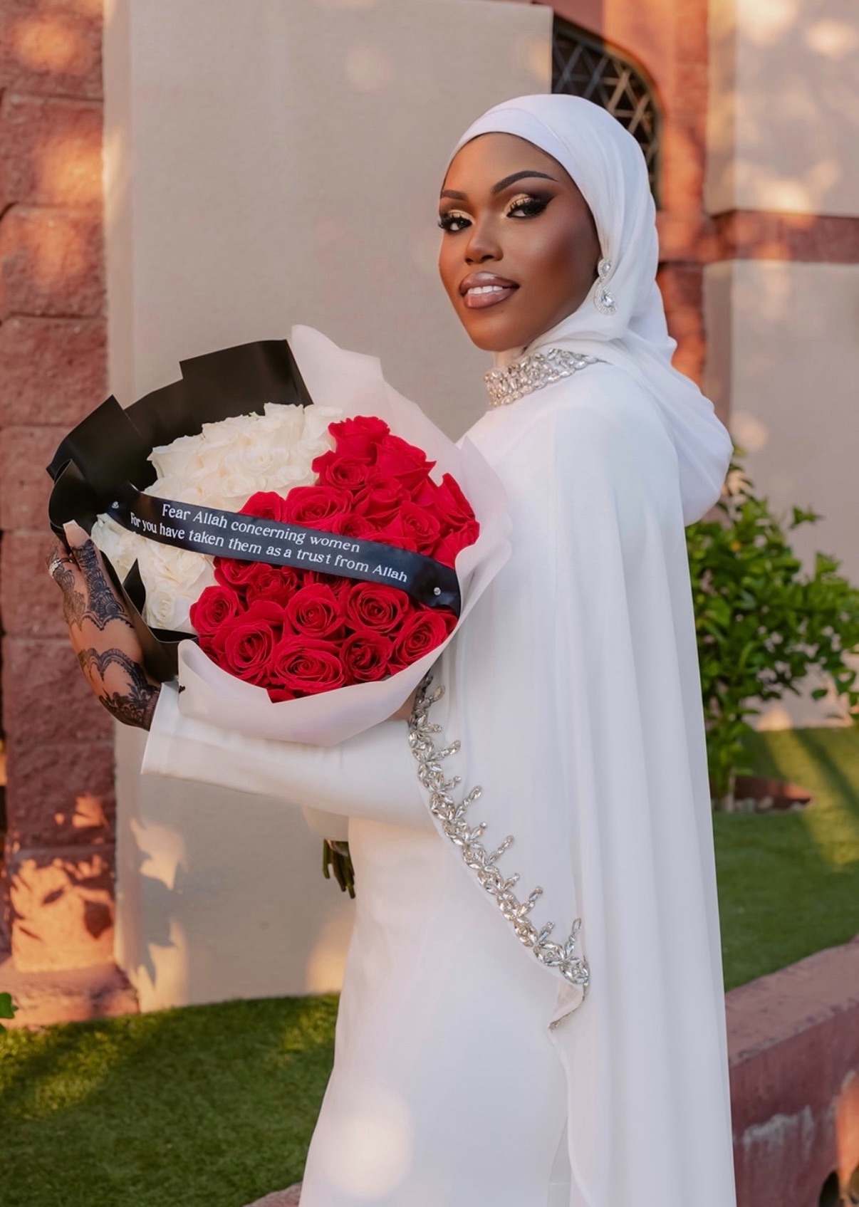 Woman in white dress and hijab holding a bouquet of red roses outdoors, smiling, with a building and greenery in background.