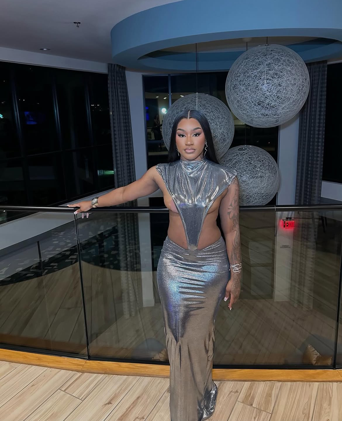 Woman in metallic dress standing indoors near glass railing with large spherical light fixtures overhead.