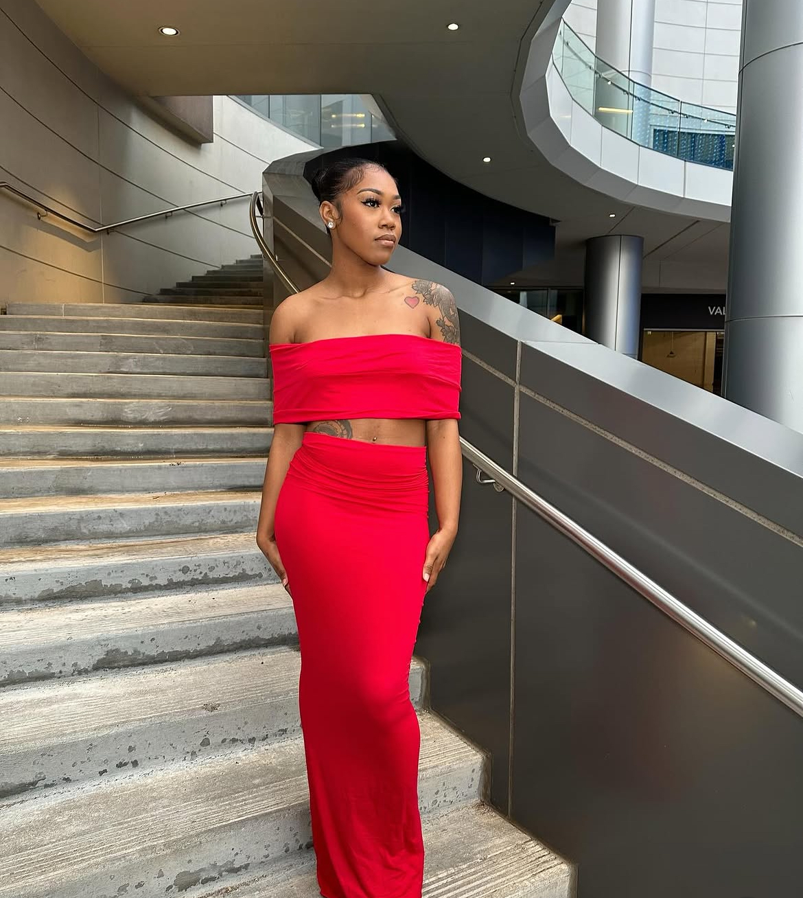 Woman in red off-shoulder dress standing on stairs in modern building interior.