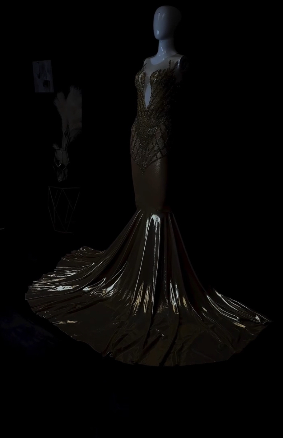 Mannequin wearing a shiny, metallic, floor-length gown with a fitted bodice and flowing train, displayed in a dark setting.