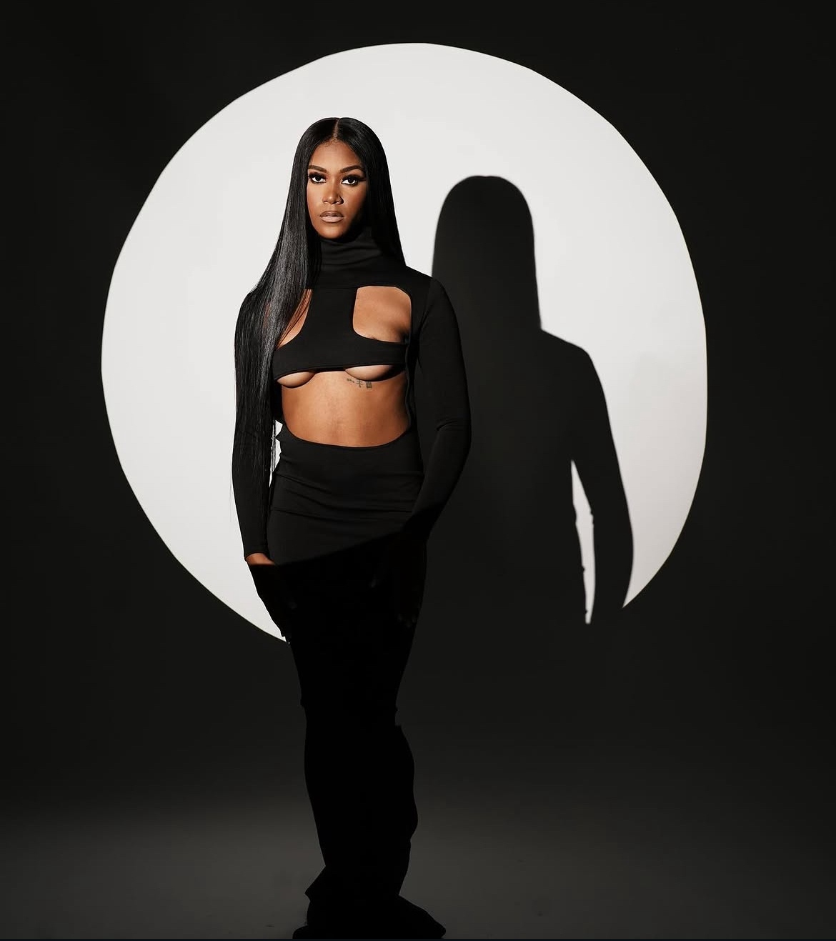 Woman with long black hair in a black cut-out outfit stands in front of a white circle on a black background.