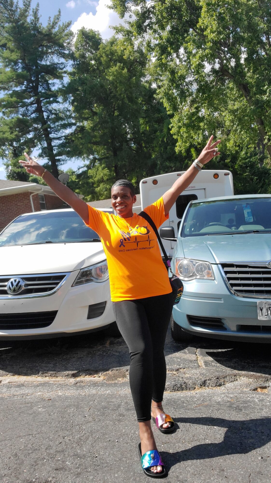 Person standing outdoors with arms raised, smiling, in front of parked cars and trees.