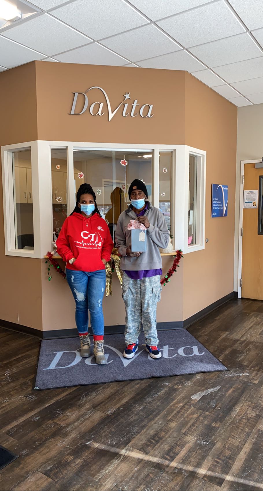 Two people wearing masks standing in front of a reception desk with a sign that says 'DaVita'.