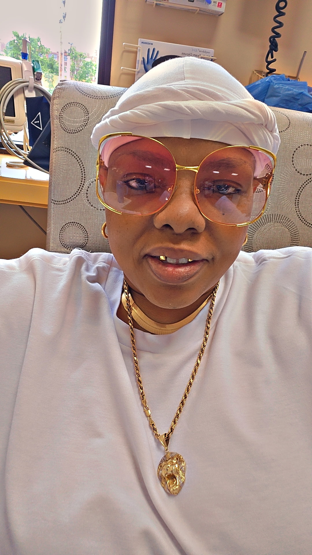 Person wearing pink sunglasses, white headwrap, white shirt, and gold jewelry, sitting in a room with a window and office supplies.