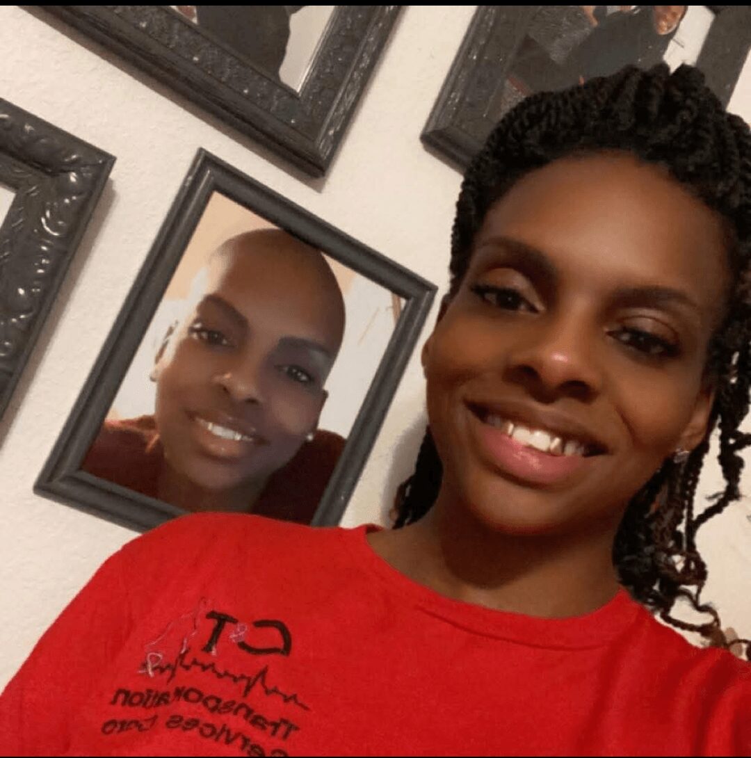 A woman with braided hair smiling, taking a selfie in front of a mirror with a framed photo of a person smiling.