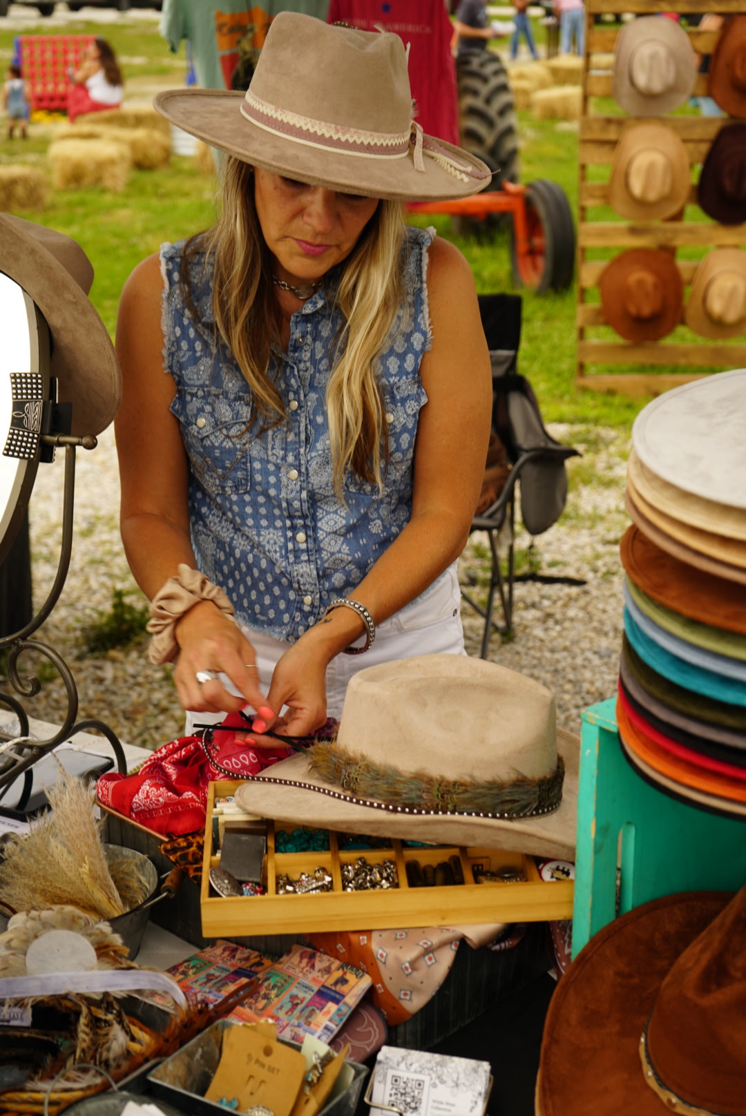 Woman browsing hats at outdoor market, wearing a wide-brimmed hat and blue patterned top, surrounded by hats and accessories.