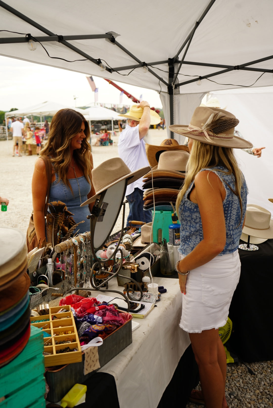 Two women at a booth under a tent, one with long hair and a hat, the other with wavy hair, surrounded by accessories and items.