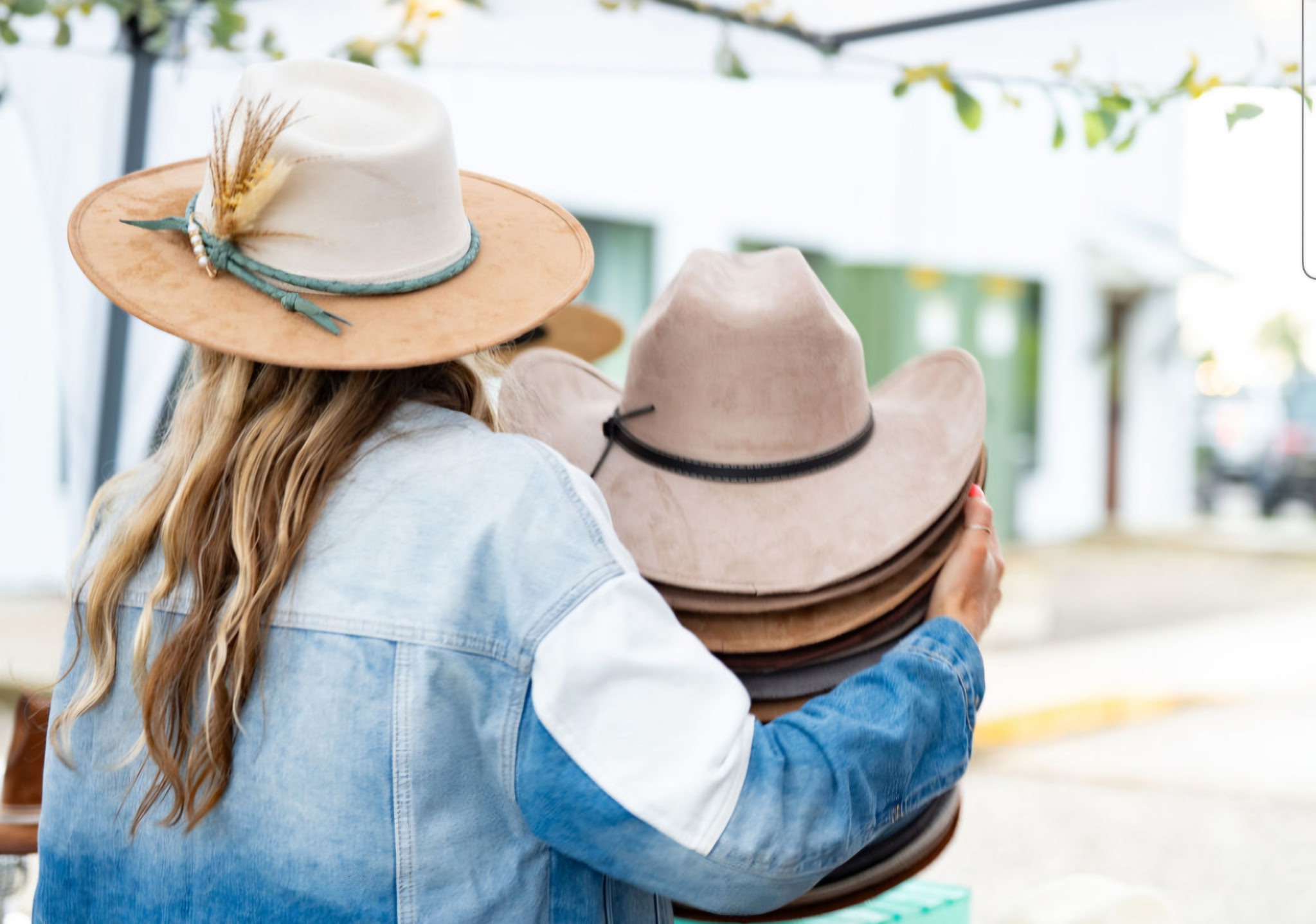 Person with long hair wearing a hat and denim jacket holding multiple hats outdoors.