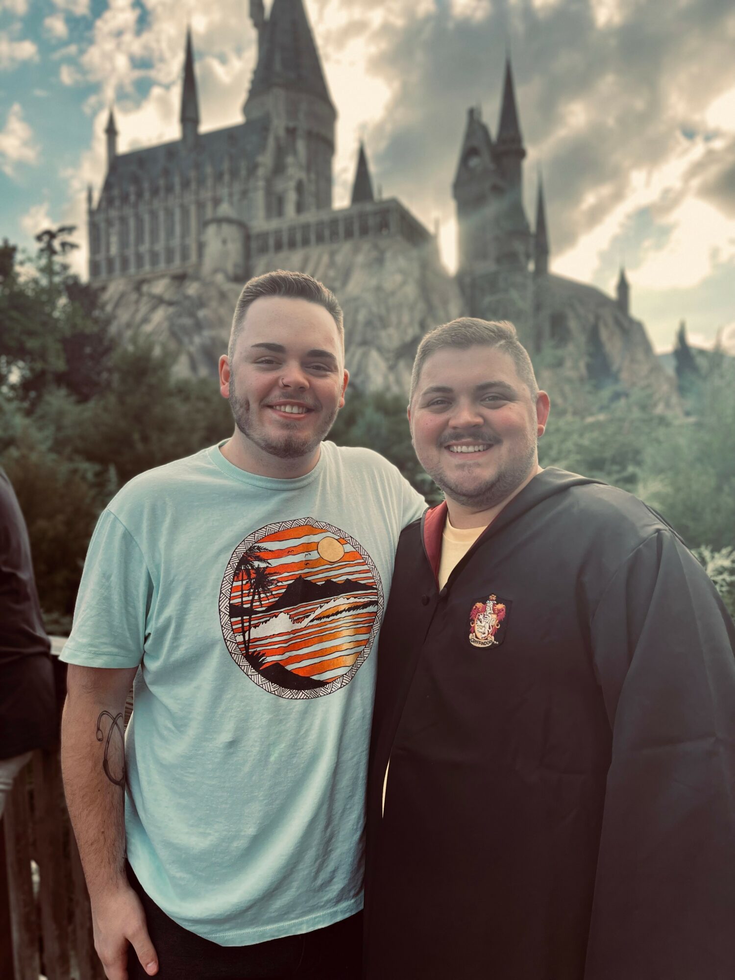 Two smiling men stand outdoors with a castle-like building in the background, cloudy sky overhead.