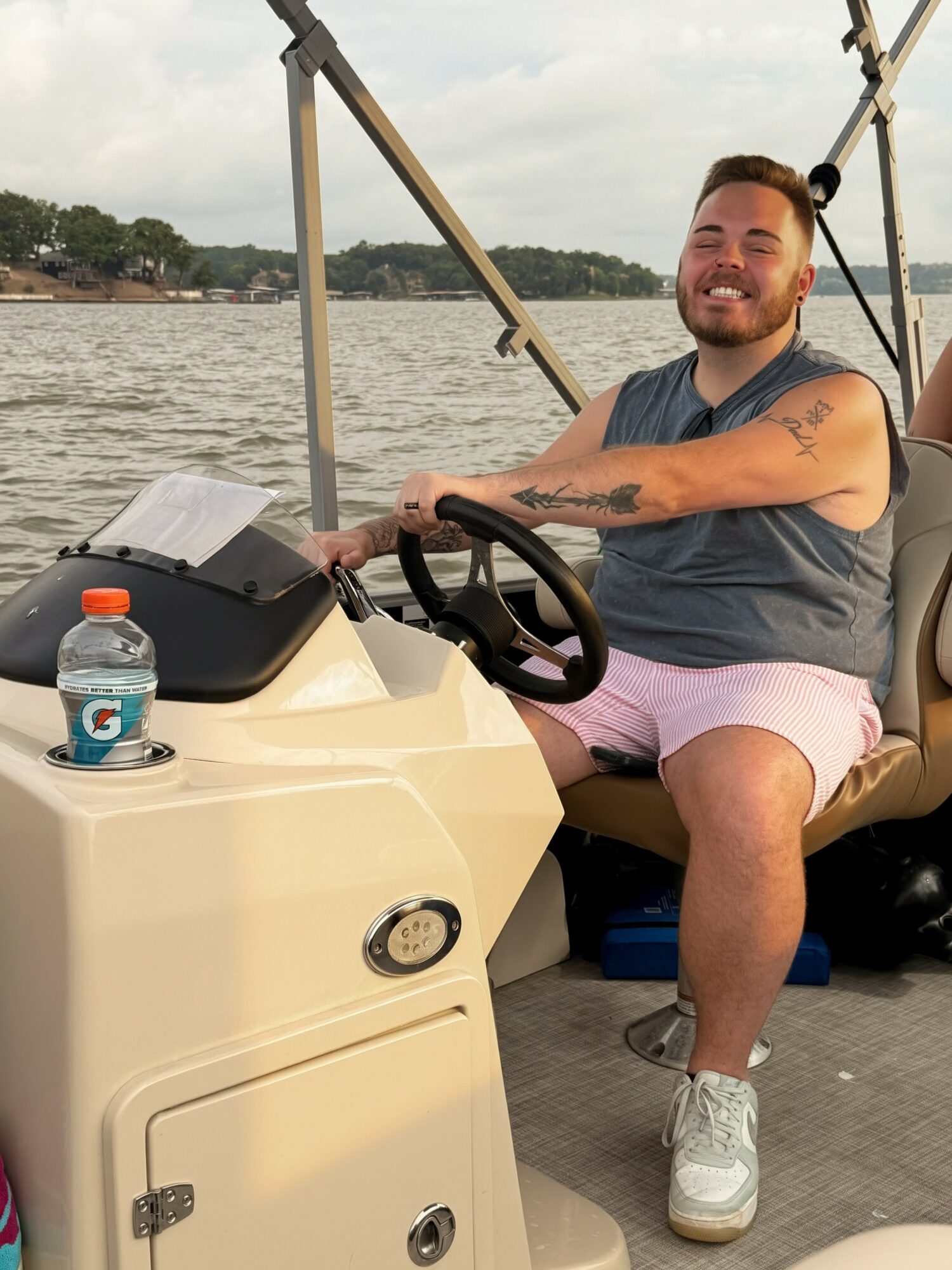 Smiling man sitting on a boat with water and trees in the background, holding the steering wheel, wearing a sleeveless shirt and shorts.