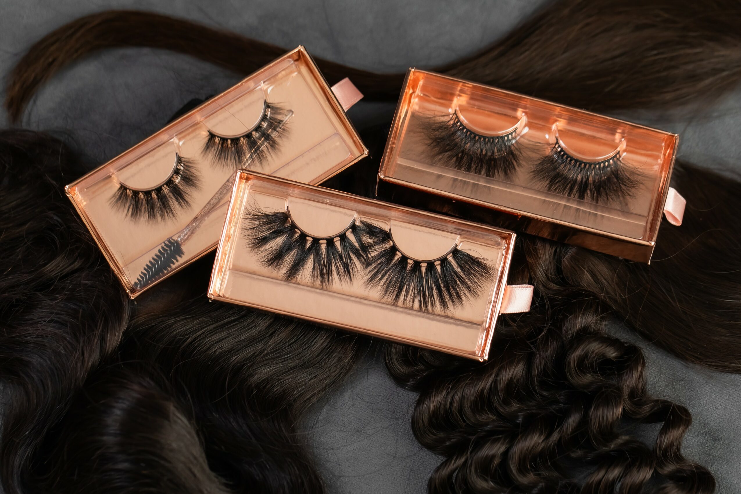 Three sets of false eyelashes in pink boxes on dark hair, with one set on a gray background, all open.