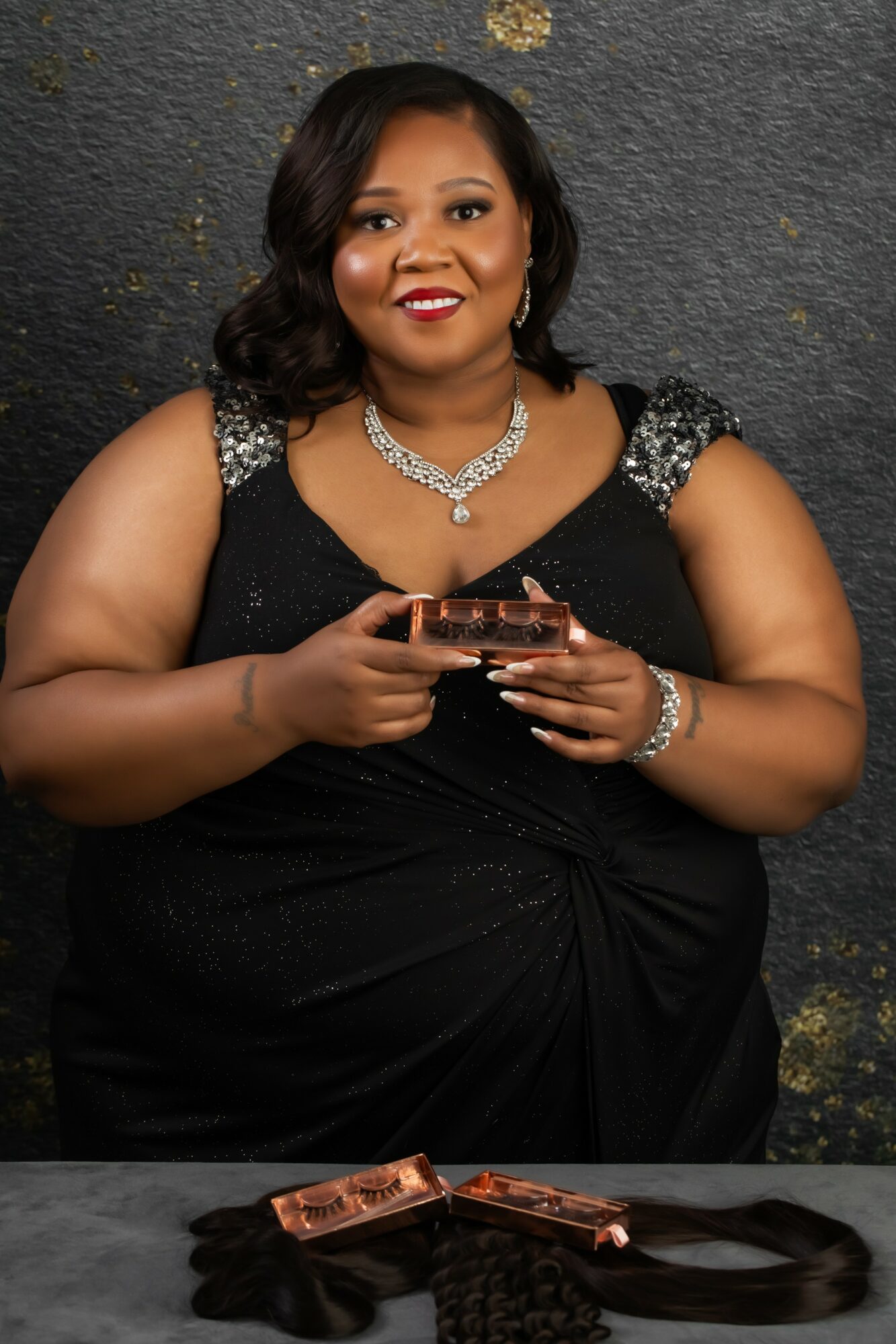 Woman in black dress with silver embellishments, holding a phone, smiling, against a dark textured background.