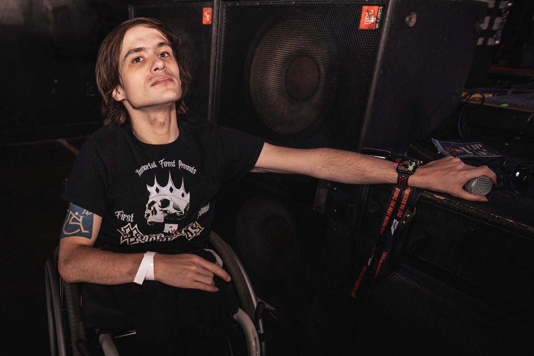 Person with long hair and a black T-shirt sitting in a wheelchair, reaching out to a DJ setup, in a dark environment.