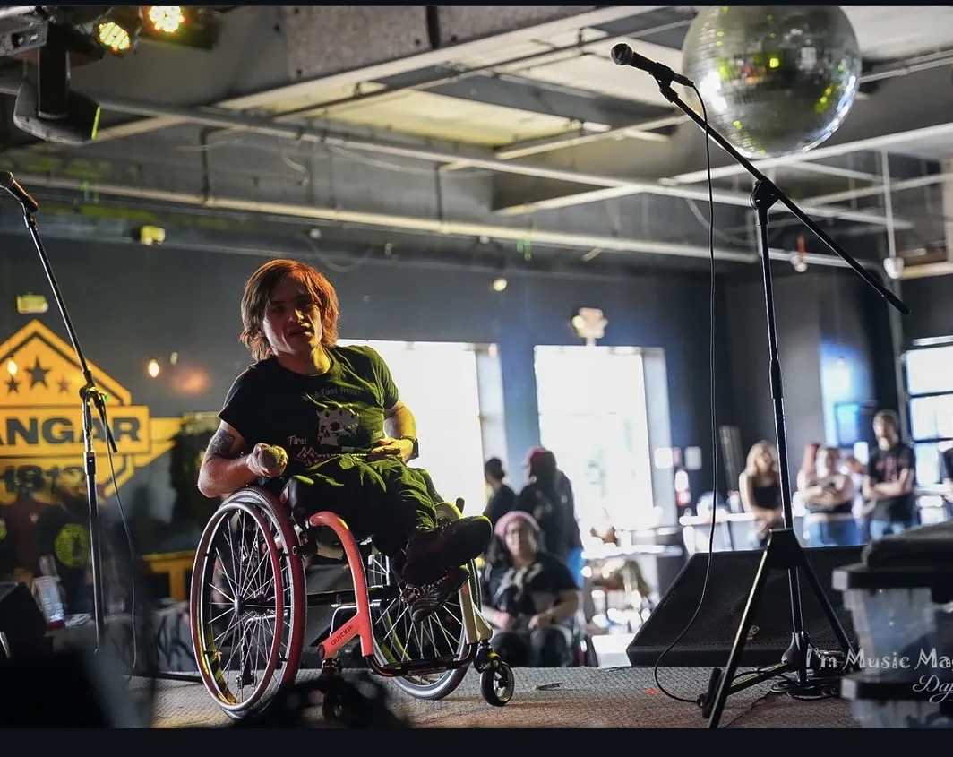 Person in a wheelchair on stage with microphones, audience in background, disco ball overhead, stage lights, and a yellow warning sign.