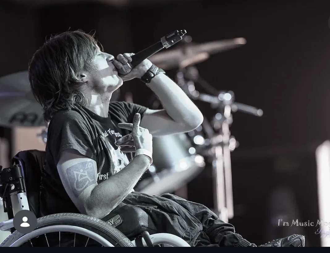 Person in a wheelchair singing into a microphone on stage with musical equipment in background.