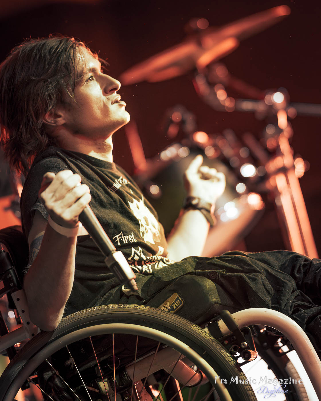 Person in a wheelchair with long dark hair, looking upward with clenched fists, on stage with drums in background.