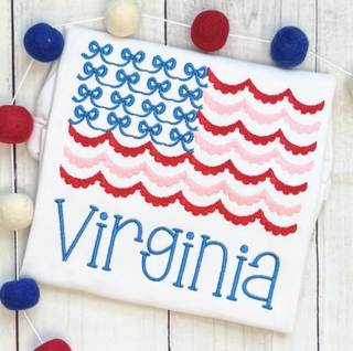 White fabric with red, pink, and blue decorative patterns and the name Virginia written in blue, surrounded by red, white, and blue pom-poms.