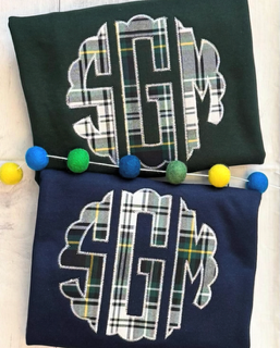 Two folded shirts with monogram designs, one green and one navy, placed on a white surface with colorful beads.