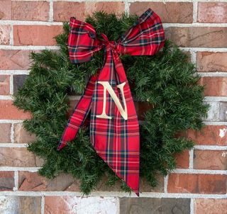 Green Christmas wreath with a red plaid bow and a gold letter N, hanging on a brick wall.