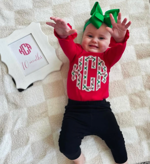 Baby girl with a green bow headband, red sweater with a holiday design, black pants, smiling with arms raised, on a beige blanket.