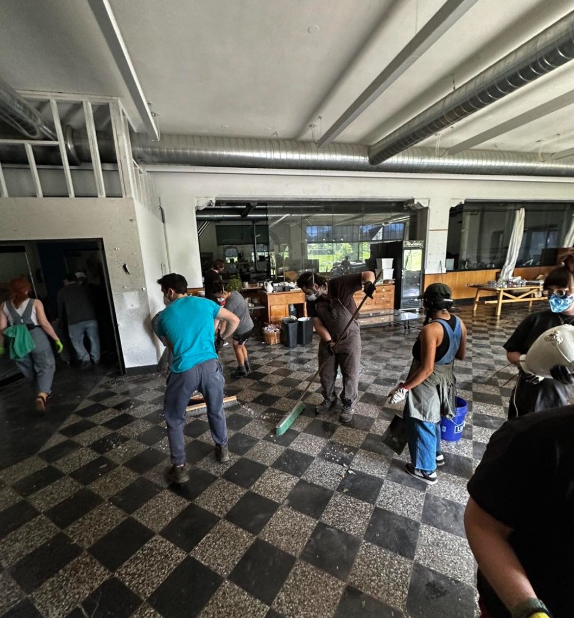 People standing and walking in an indoor space with checkered flooring and large windows, some wearing masks.