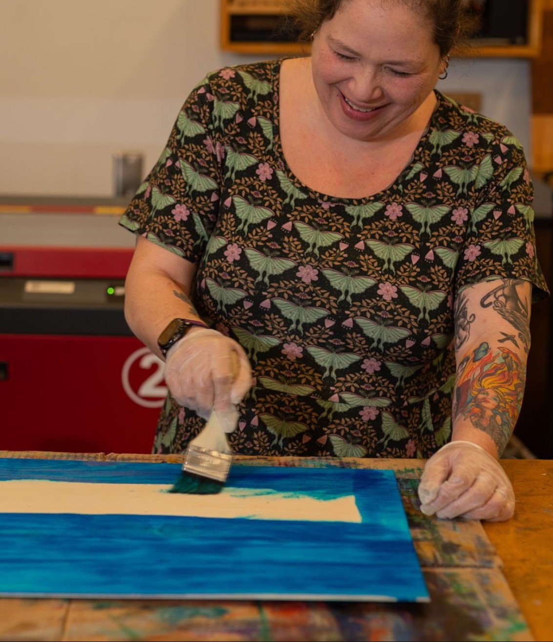 Woman smiling and painting a blue border on a canvas, wearing a floral shirt and gloves, in an indoor setting.