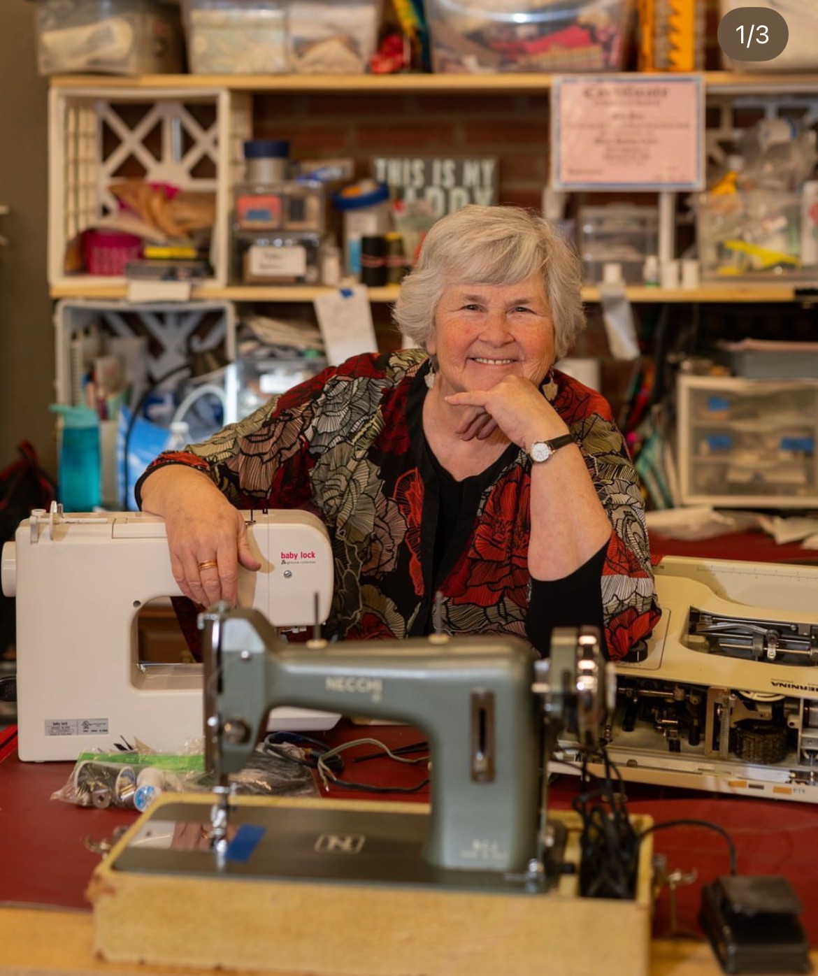 Older woman smiling at sewing machine in craft room, surrounded by supplies and shelves.