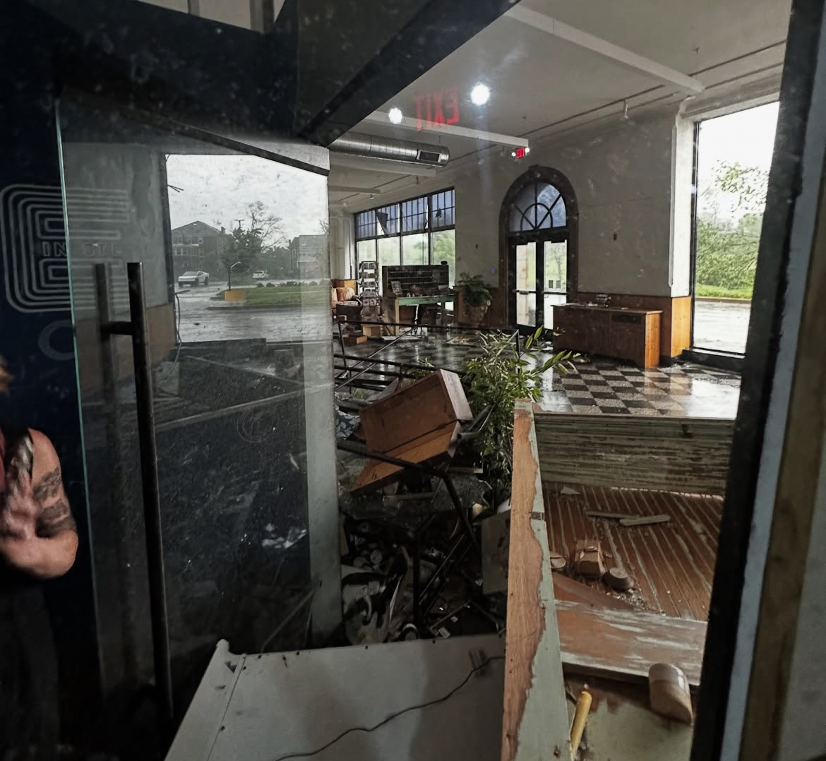Interior of a building with damaged furniture and debris, large windows, and a checkered floor, seen through a glass door.