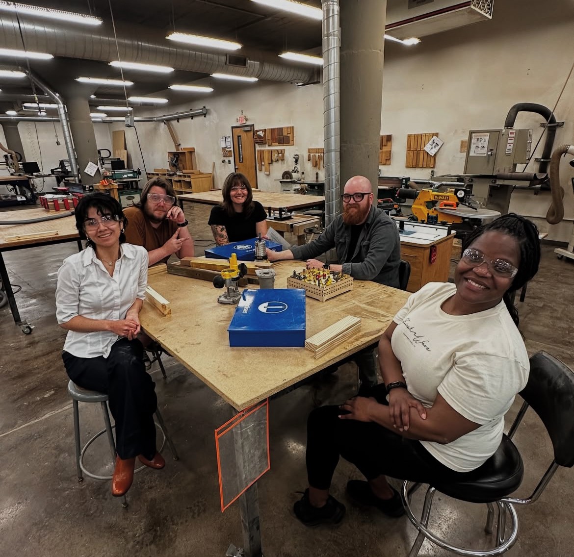 Five people sitting around a table in a workshop, smiling, with woodworking tools and equipment in the background.