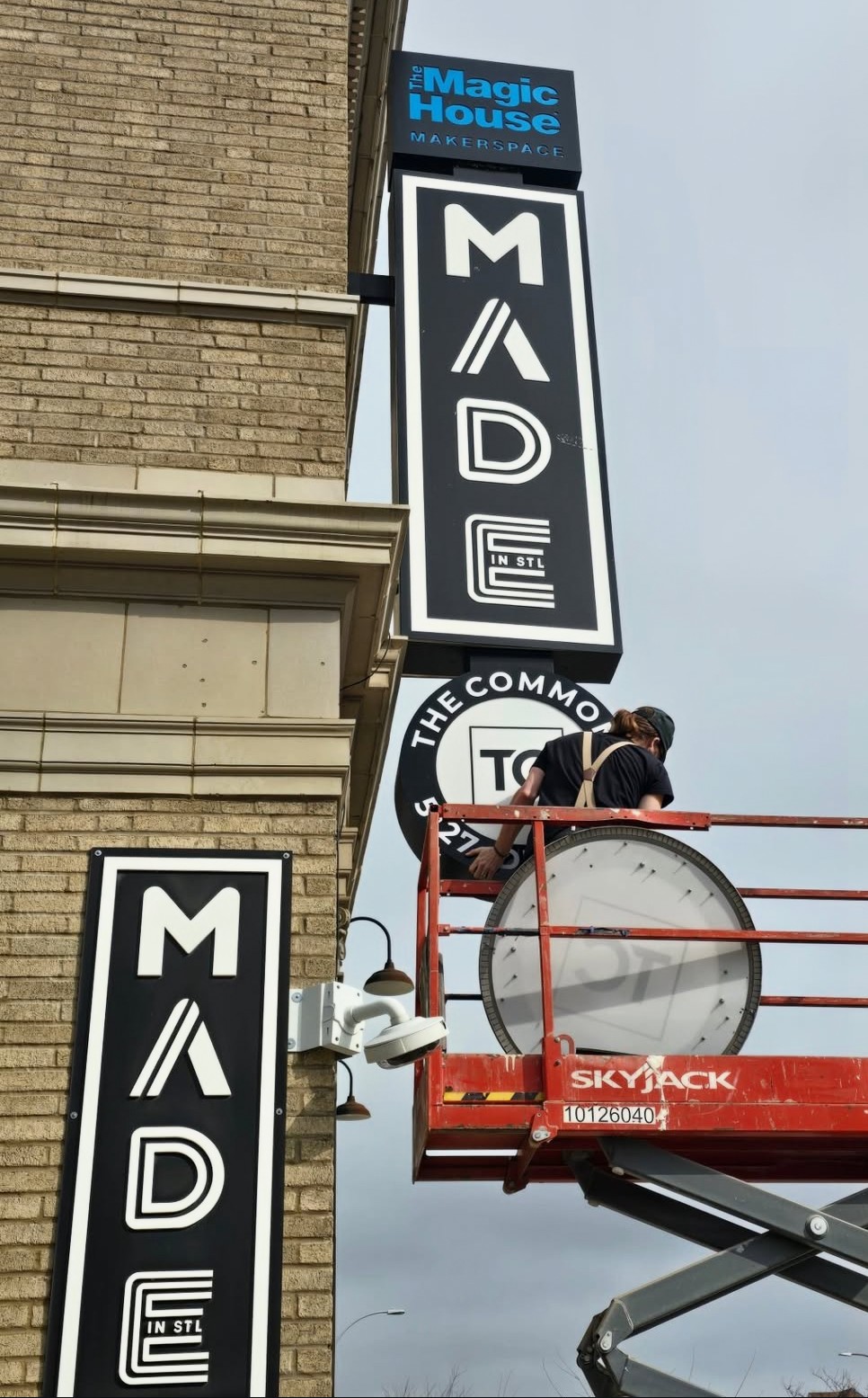 Two vertical signs for 'MAD E' and a blue sign for 'Magic House' on a brick building, with a person on a lift working on the signs.