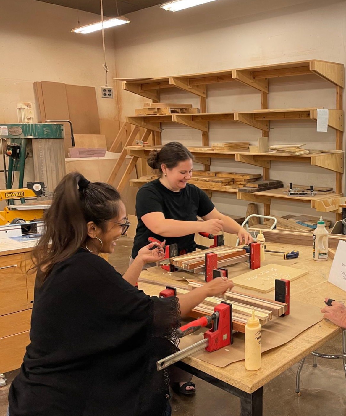 Two women working on woodworking projects at a workshop table with wood pieces and clamps.
