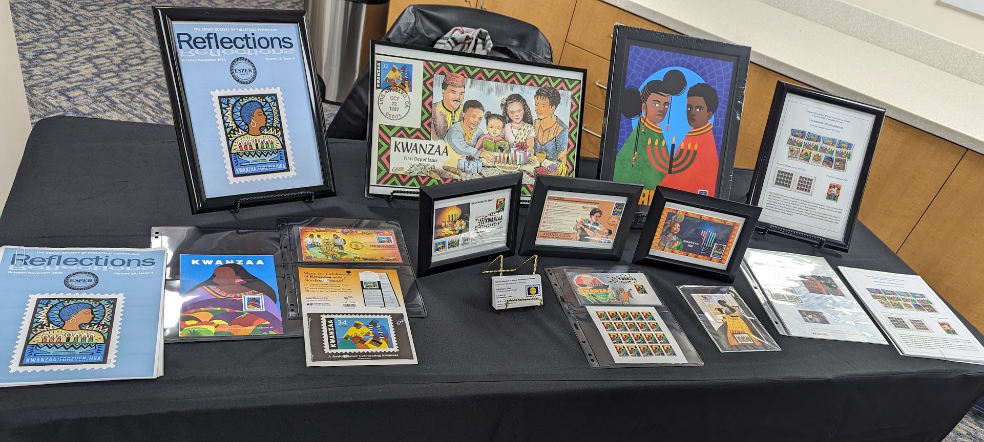 Display table with framed artwork, photographs, and printed materials arranged on a black tablecloth.
