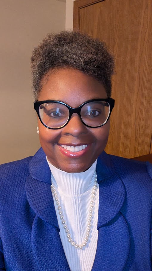 Woman with short curly hair, glasses, wearing a blue blazer, white turtleneck, and pearl necklace, smiling.