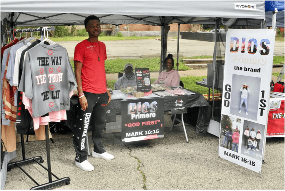 Young man standing next to a booth with religious posters and clothing, outdoors on a cloudy day.