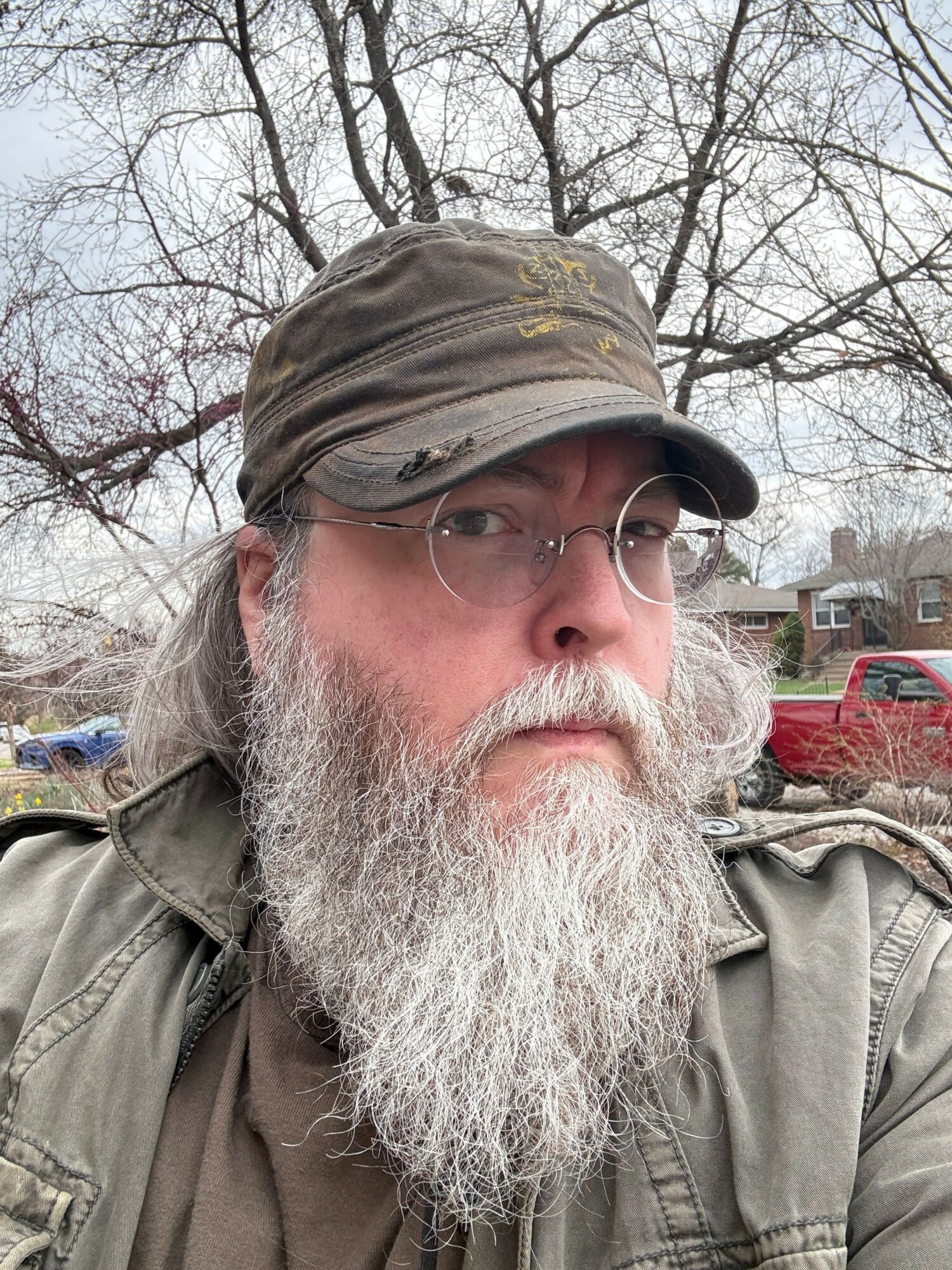Older man with a long white beard, glasses, and a cap outdoors, trees and buildings in background.