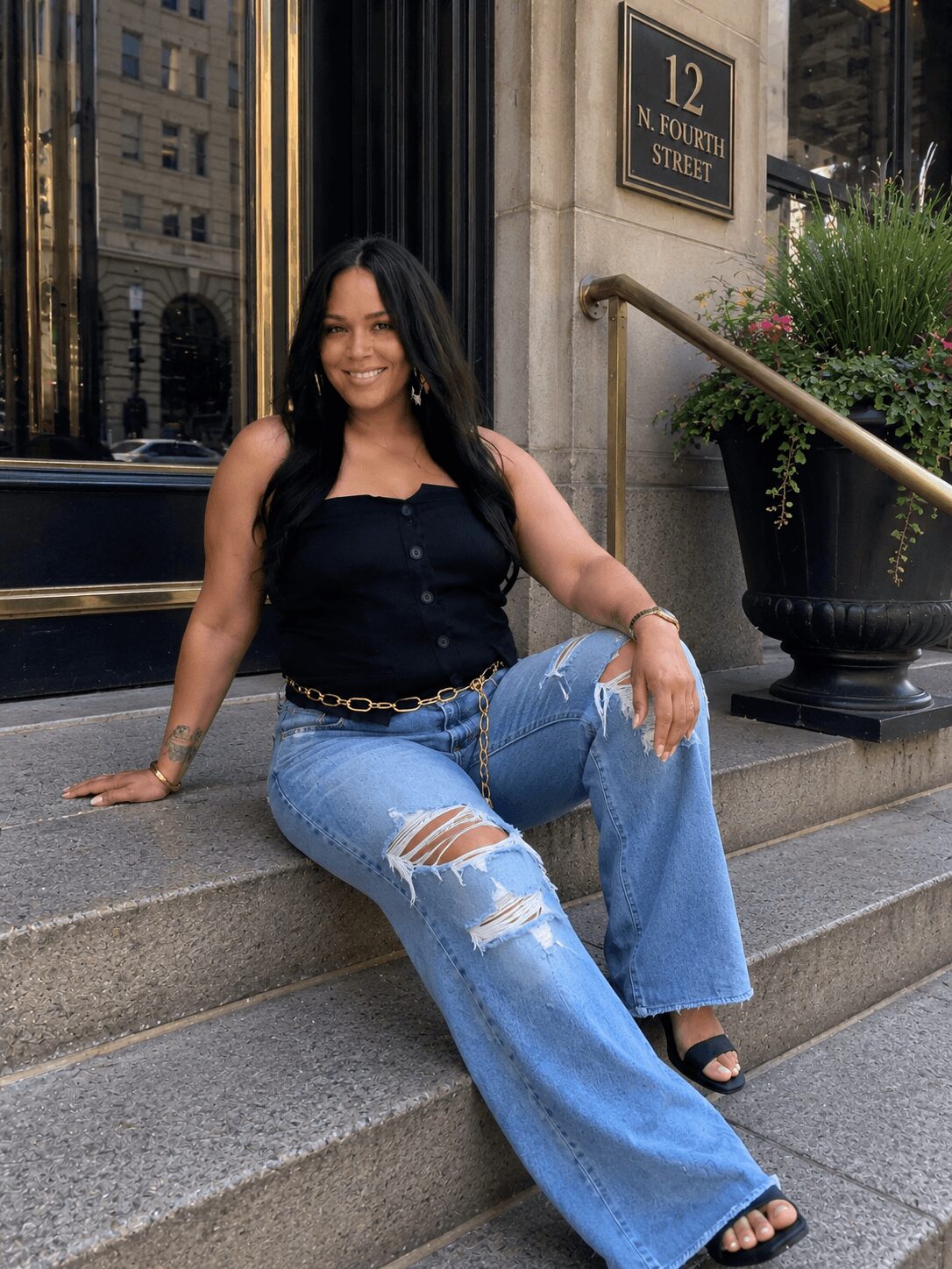 Woman sitting on steps outside building, smiling, wearing black top, ripped jeans, and sandals, with a large potted plant nearby.