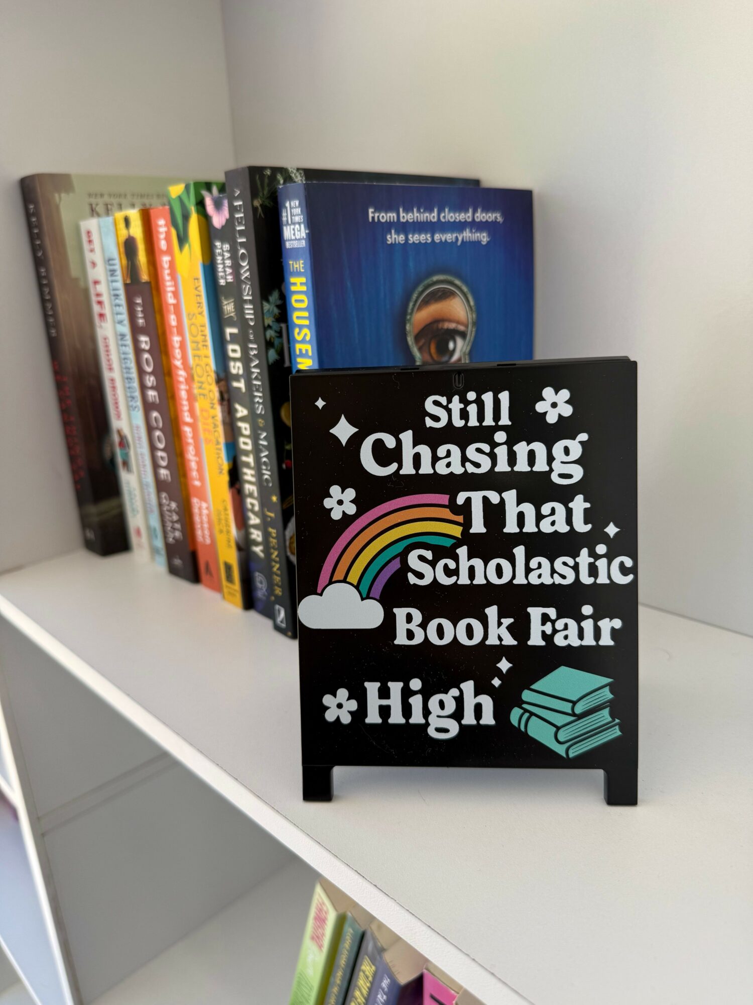 Books stacked on a white shelf with a black sign that reads 'Still Chasing That School Bell, Fair High' with rainbow and cloud illustrations.
