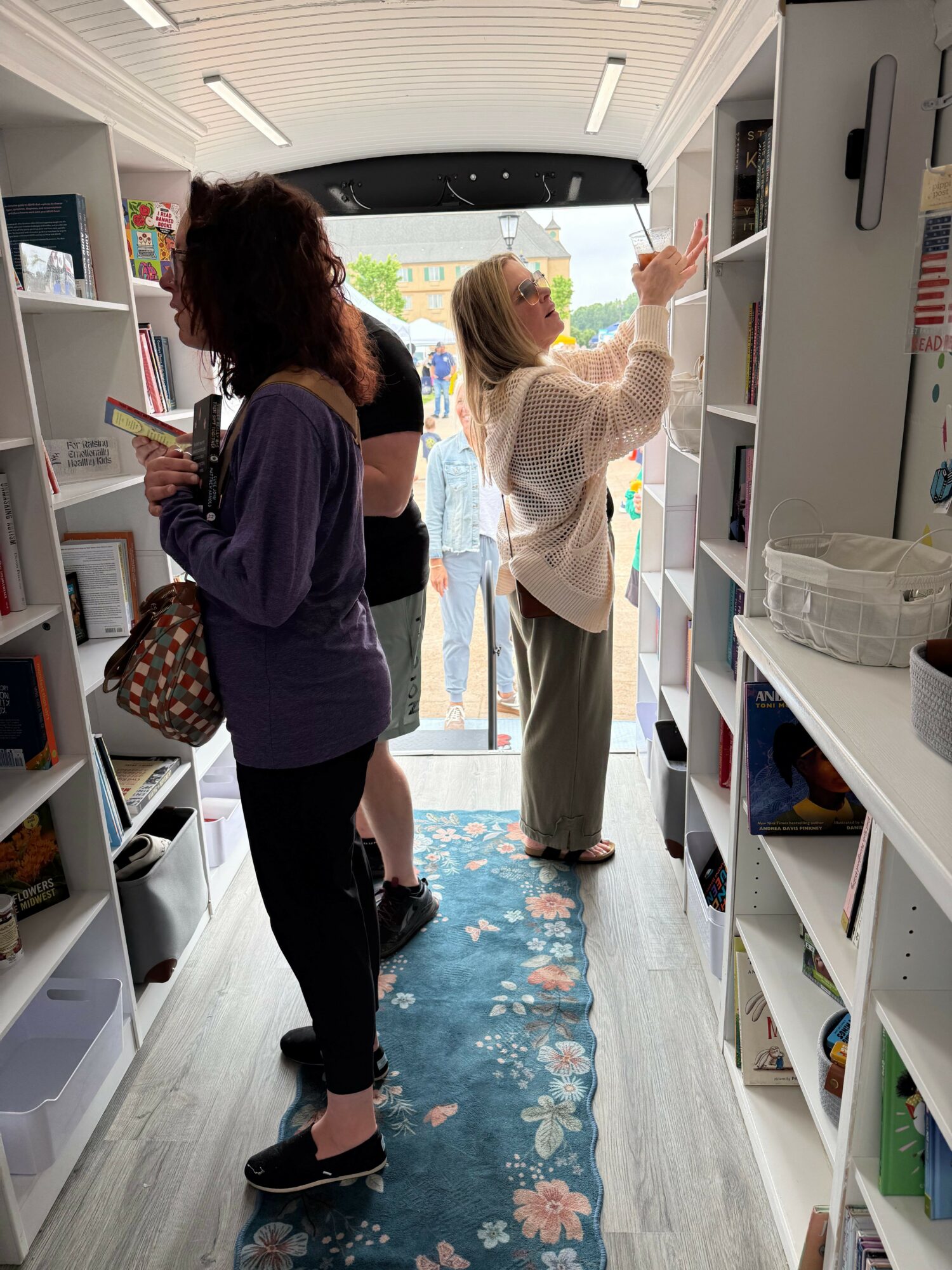 Two women stand inside a bookstore near a large window, browsing books on shelves, with a blue floral rug on the floor.