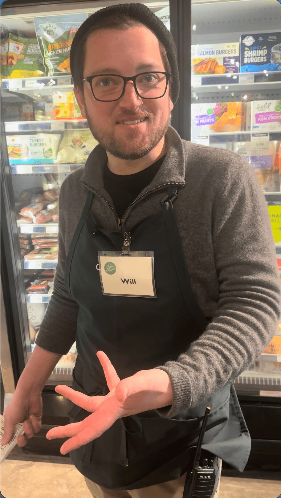 Person with glasses and a name tag smiling in a store, holding up four fingers, with refrigerated products in the background.